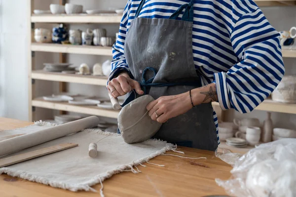 Pottery making process. Woman ceramist in apron creating crafted and artistic objects, cropped photo of female potter using clay modeling tool while standing at workplace in studio or workshop