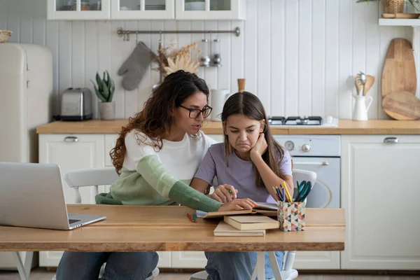 Attentive caring woman helps daughter prepare for exams for transfer to new school. Dissatisfied girl teenager sits with mother at kitchen table unwilling to study and do extracurricular work