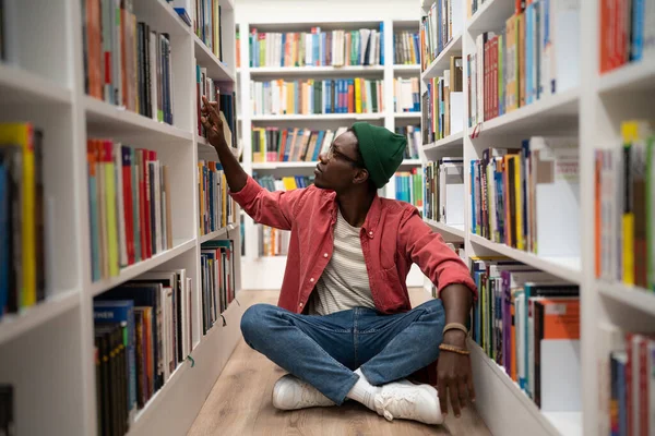 Self education. Young African American guy sits on floor in library between bookshelves, spending time at bookstore, searching for book, black male student preparing for college test. World book day