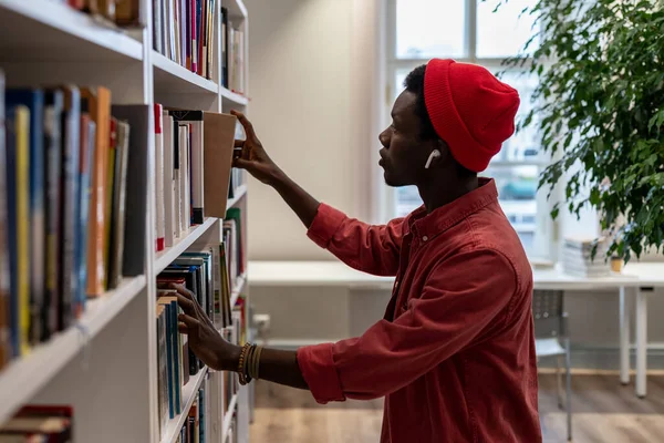 Thoughtful African American student man searching materials for educational research in college library. Young black hipster guy choosing book for reading in bookstore. Literature and education