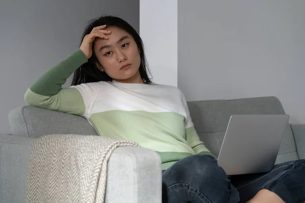 stock image Procrastinating pensive Asian female blogger sits on sofa with laptop on lap and looking away dejectedly. Unmotivated Japanese freelance woman is distracted from work due to lack of inspiration 