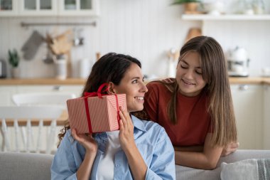 Happy festive family of woman and teenage girl giving mother gift box on holiday or birthday. Kind positive daughter stands behind nanny sits on sofa in living room, intrigued by unexpected president