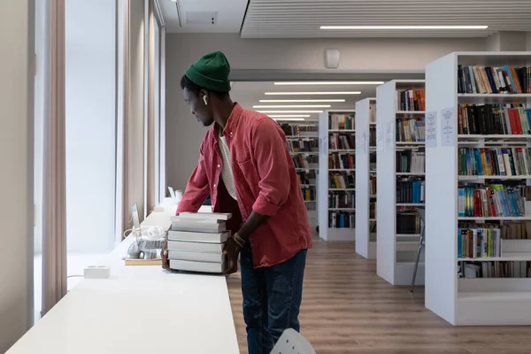 アフリカ系アメリカ人男性は大学のための科学報告書を準備する図書室に立っている ヘッドフォンのカジュアル自給黒人男性は書店の建物でノートパソコンの横にテーブルの上に本を置きます — ストック写真