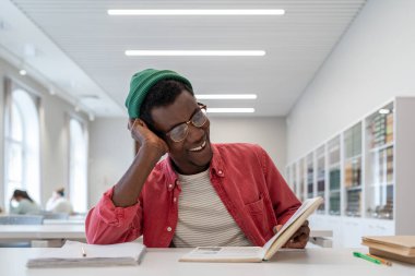 Laughing cheerful African American man reading book sits at table during break between classes in college or high school. Carefree happy black guy enjoys favorite hobby reading fiction in library  clipart