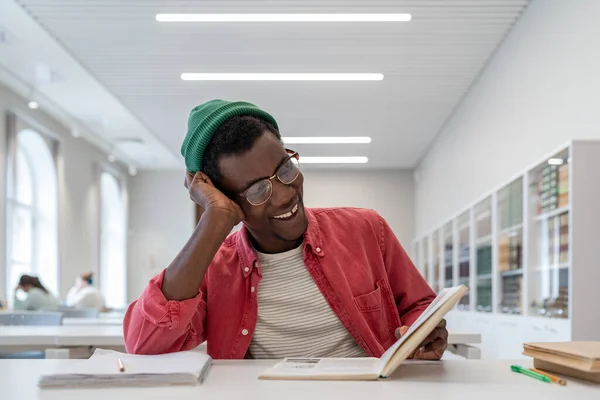 Laughing cheerful African American man reading book sits at table during break between classes in college or high school. Carefree happy black guy enjoys favorite hobby reading fiction in library