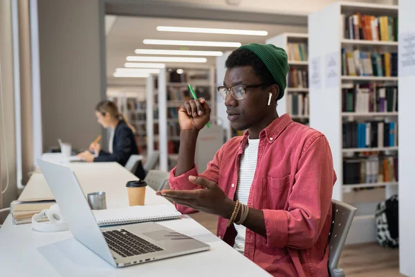 スマートフォーカシングアフリカ系アメリカ人男性は 職場の同僚とビデオ通話で話す公共図書館のノートパソコンとテーブルに座っています オンライン講義を見ている若い男の学生は難しい試験の準備 — ストック写真