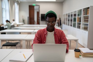 Focused Black man in glasses sits at library table with laptop, studying online in modern classroom. Concentrated African American student guy using internet in reading room, preparing for exam. 