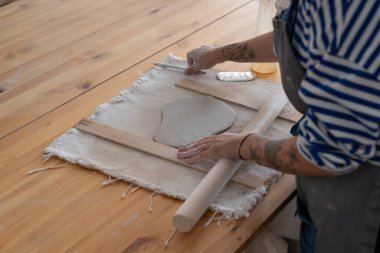 Woman potter artist in apron use wooden rolling pin to roll out clay making handmade utensils. Process of creating dishes by professional ceramics specialist in workshop with table made of planks