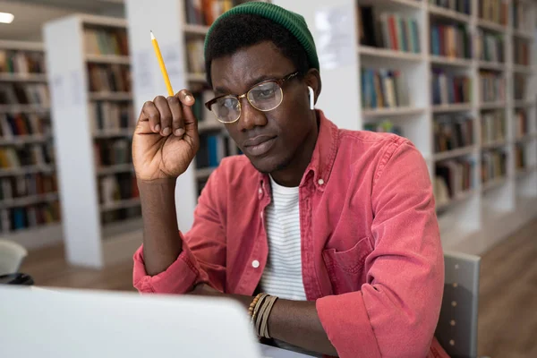 Focused African American student guy wearing glasses and wireless earbuds sitting in library looking at laptop screen, watching online lesson, using webinar platform in education. E-learning concept