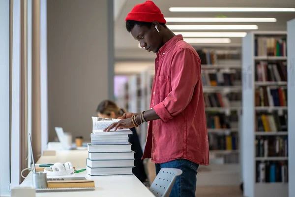 アフリカ系アメリカ人の学生の男の机の上に本の山で 論文や研究プロジェクトに取り組んで 本やその他の情報源を検索するためにライブラリリソースを使用して立って — ストック写真
