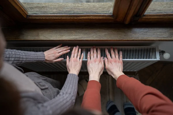 Top view of couple warming hands above radiator in cold house, cropped photo. People keeping warm in winter at home. Heating season and energy crisis concept