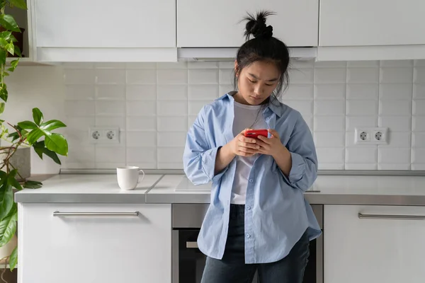 stock image Concentrated emotionless Asian woman typing text on phone standing in kitchen with glass coffee. Carried away Korean girl chatting on social media is digitally addicted spending lot time on internet