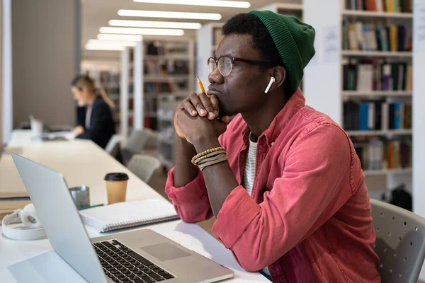 Thoughtful black male student wearing wireless earbuds sitting at desk with laptop in library, listening English audio lessons. African American student guy looking out window during online class