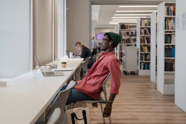 Yorgun Afrika kökenli Amerikalı serbest çalışan bir erkek dizüstü bilgisayarla masada oturuyor ve kütüphanede uzaktan çalışıyor. Mutsuz zenci öğrenci kendini çalışmaktan bunalmış hissediyor. Çevrimiçi öğrenmede motivasyon eksikliği