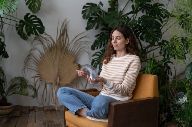 Relaxed calm woman playing with spring toy for stress relief sit crossed legs in armchair. Female relieve stressful tension with stretchable spring has break from work at home office in indoor garden clipart