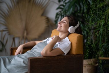 Relaxed girl lies surrounded by exotic green plants in the city botanical garden in wireless headphones. Smiling woman resting after working day listening to music, interesting podcasts in earphones.