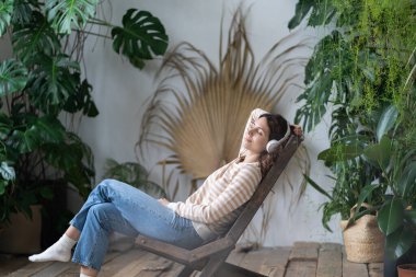 Young woman with closed eyes and put hand back wearing wireless headphones, rest in work break, sits on wooden chair in urban jungle garden with monstera, enjoy good relaxed music. Stress free concept