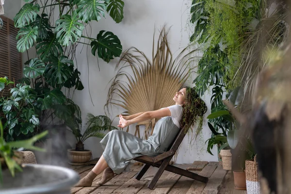 Health and well-being benefits of plants. Young happy dreamy italian woman in dress relaxing in tropical resort-style garden at home, relaxed spanish female gardener resting after work in greenhouse