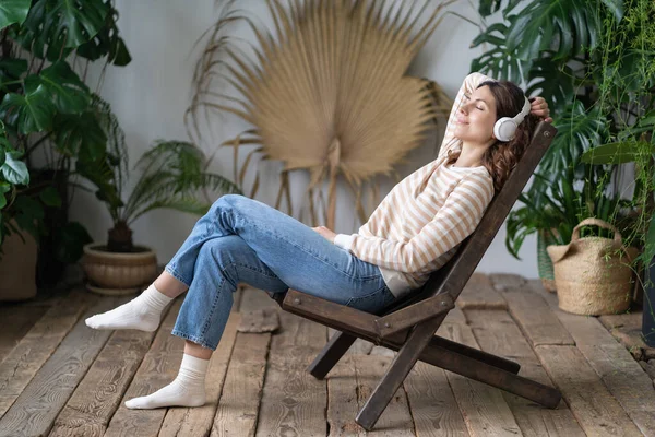 stock image Calm woman sitting in armchair in cozy greenhouse, dreaming, resting, listen to music on wireless headphones. Female wear earphones, closed eyes and relaxing surrounded by houseplants in living room.