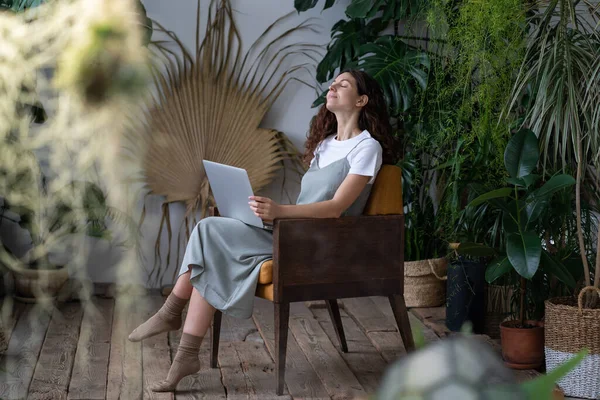 Relaxed woman gardener with closed eyes holding laptop sitting in chair surrounded by various tropical plants, full-length. Female online plant shop owner enjoying remote work in home garden