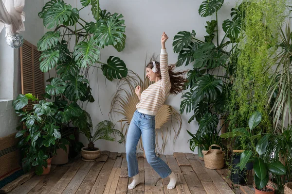 stock image Positive young italian woman in wireless headphones listening to music with pleasure, enjoying the moment, relaxing, dancing on wooden floor in cozy home garden with monstera and tropical plants. 