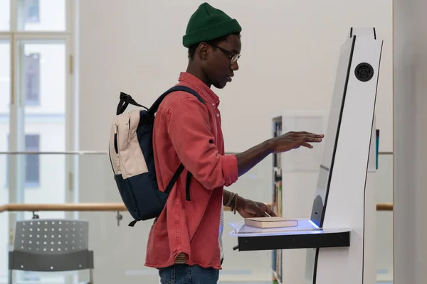 図書を借りたり返却するためにセルフサービス端末を使用してライブラリに立っているバックパックを持つ若いアフリカの学生の男 文献閲覧カタログを検索 タッチスクリーン自己チェックアウトキオスク — ストック写真