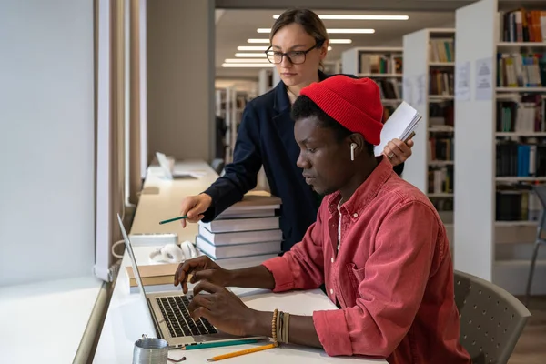 図書館の机に座っているアフリカの男に何かを説明しながら ノートパソコンの画面を指している若い女性教授 大学の入学試験のための学生を準備教師 教育における技術 — ストック写真