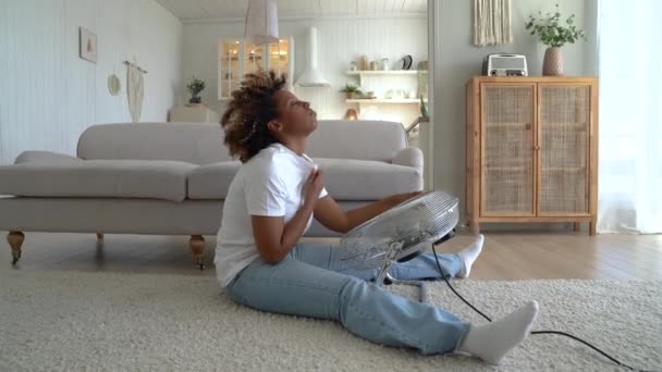 過熱した家 若い夏の暑さから疲れアフリカ系アメリカ人女性の床に電気ファンを使用して座っている 不幸な黒の女の子が熱い閃光に苦しんでクールダウンする ホルモンの不均衡 — ストック動画