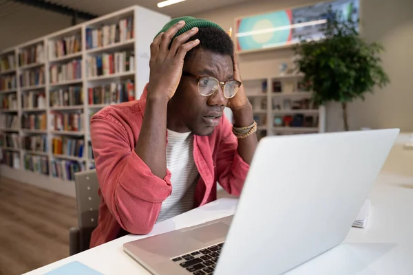 ガラスの衝撃を受けたアフリカ系アメリカ人の学生は 図書館のテーブルに座ってノートパソコンの画面のクラッチヘッドを見て混乱しているように感じます プログラムのエラーから複雑化した黒男を混乱させると デバイスのマルウェアを取得します — ストック写真