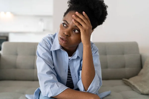 Flustered African American woman very nervous sighing and touching face with hands sits on couch of home. Young black girl nervous, thinking, feeling fear after wrong decision or problems at work.
