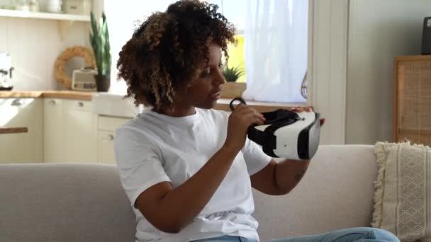 虚拟现实的初步经验 年轻的非洲裔美国女人坐在沙发上 手持Vr护目镜 探索数字世界 兴奋的黑人女孩测试新的高科技礼物Ar装置 — 图库视频影像