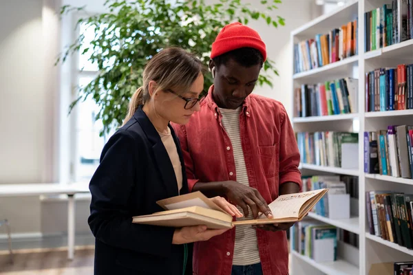 アフリカ系アメリカ人の学生は 大学の本棚の近くでプロジェクトパートナーに試験準備書に記載されている情報を示しています 大学図書館でパズルカップルが割り当てで忙しい見て — ストック写真