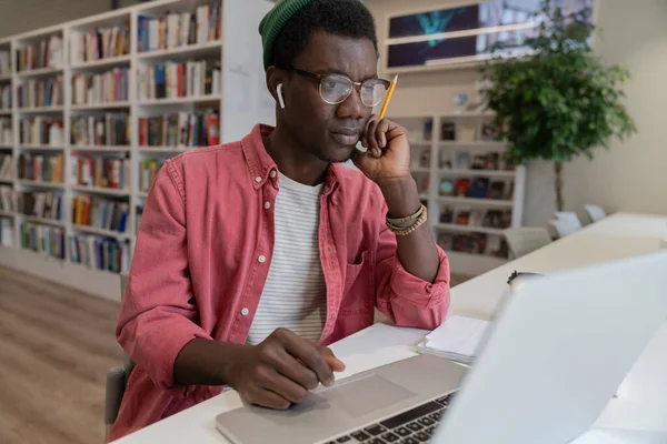 重点是在校园图书馆中对黑人学生进行新观念的探索 专心致志的非洲裔美国自由职业者拿着铅笔 用笔记本电脑坐在工作桌旁 沉思着任务的解决方案 — 图库照片