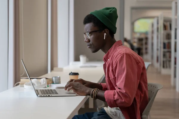 专注的非裔美国人在公共图书馆的全景窗口附近的笔记本电脑上留言 以黑人为焦点的人在网上交流 发邮件 在电脑上远程工作或学习 — 图库照片