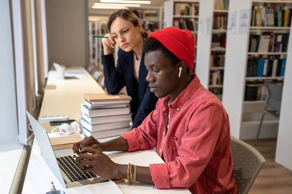 黒人学生の男がラップトップを使用してオンラインタスクを解決するのを助けるプロの集中教師の女性メンターを動揺 大学図書館での挑戦的な研究プロジェクトに関する多民族の困惑学生のチームワーク — ストック写真