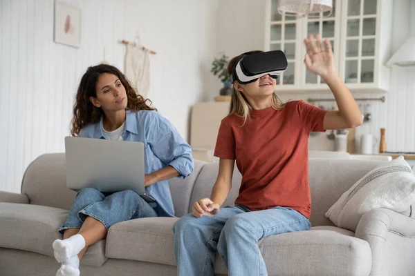 stock image Interested teenage girl uses VR headset to visit metaverse or watch 3D virtual reality movies from comfort home. Progressive European schoolgirl waving hands sits on sofa near mother woman with laptop