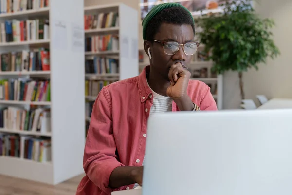 図書館の職場のテーブルでノートパソコンで働く眼鏡のアフリカ系アメリカ人のヒップスターフリーランスの男性に焦点を当て Itオンラインスタートアッププロジェクトについて考えています 集中黒男学生はタスクを解決します — ストック写真