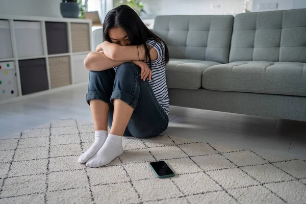 Upset unhappy sad asian woman sitting near smartphone on floor at home. Feeling lonely depressed heartbroken Korean girl sits crossed arms on knees looks on cellphone wait call thinks about problems
