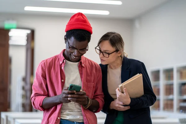 満足笑顔アフリカ系アメリカ人の学生は スマートフォンの画面上の教科書とメガネで深刻な包括的な白人同級生の女の子を表示します新しい作成されたアプリ ウイルスビデオネットワーク 研究休憩面白い投稿 — ストック写真