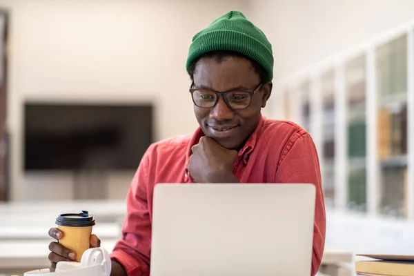 紙コップの感じでコーヒーと普及したアフリカ系アメリカ人男性はテスト結果に満足し プロジェクトの仕事を誇りに思って あごをリラックス保持します 笑顔の黒人男を熟考フリーランスはノートパソコンの画面を見て — ストック写真