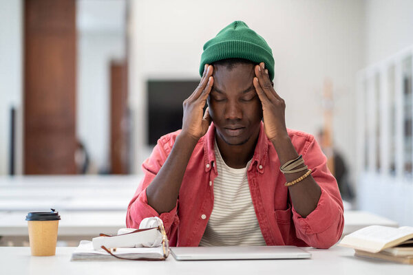 Stressed tired African American student man touching temples have headache after long time preparing for test in college library. Frustrated black hipster male feels nervous afraid of exam failure. 