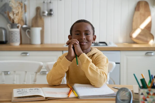 Happy Black schoolboy learning at home with textbook. Smiling African American kid boy sits at desk with pencil look at camera enjoy doing lessons in living room. Son studies while parents at work