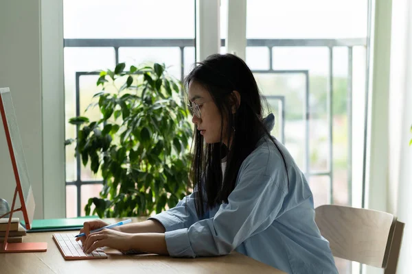 以沉思的亚洲女学生为重点 利用电脑在靠窗的舒适家中学习 严肃的年轻韩国女性戴着眼镜坐在桌旁看着键盘聊天写电子邮件发短信 — 图库照片