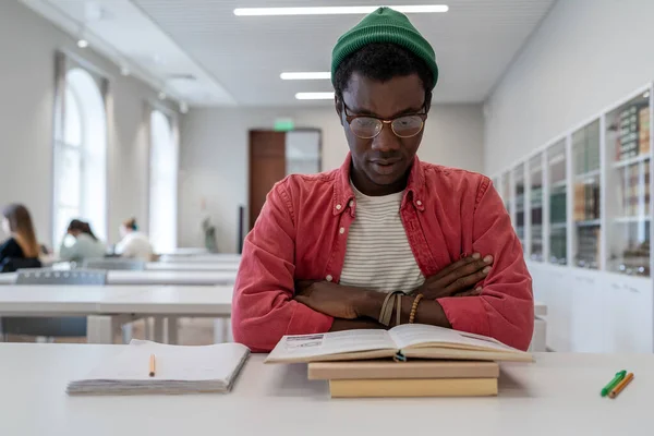 Book focused African American student guy reads study information with arms crossed in preparation for difficult exam in university library. Thoughtful man in eyeglasses reads textbook difficult task