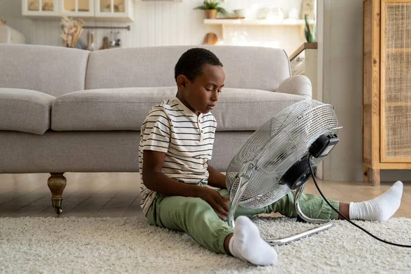 好奇的非洲裔美国男孩坐在地板上触摸电扇 享受凉爽的新鲜空气 同时在炎热的一天呆在家里 孩子和夏天的热 孩子的电安全 — 图库照片