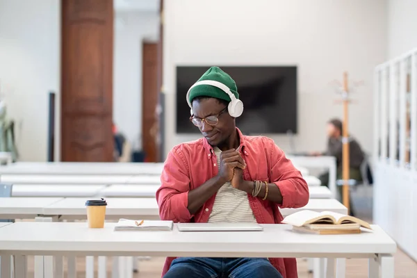 ヘッドフォンのヤーンの眠い黒人学生の男は勉強に焦点を当てることができない大学の教室の机に座っている間伸びます ノートパソコンの男との長い仕事から疲れて休憩を取るしようと残りの膝のヤシ — ストック写真