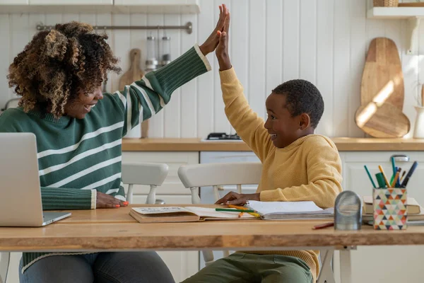 Joyful friendly mother and biracial son giving high five sitting at kitchen table celebrating success in study. Effective good cooperation result teamwork learn motivation in homeschooling with parent