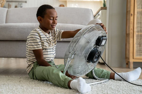 快乐的非洲男孩在客厅里独自坐在地板上 耳目一新 在炎热的夏季 使用螺旋桨的复古通风机装置给学龄前黑人儿童带来了强烈的新鲜空气打击 — 图库照片