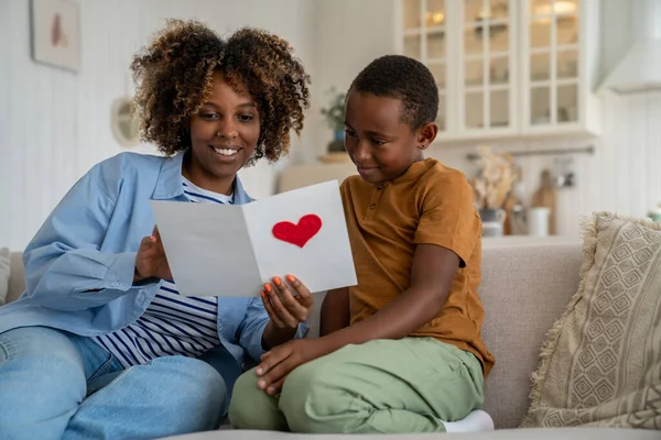 Black mum reading wishes on birthday, valentine or mother day from handmade greeting card surprised with congratulations from little son. Happy African mom and kid sitting on sofa at home together