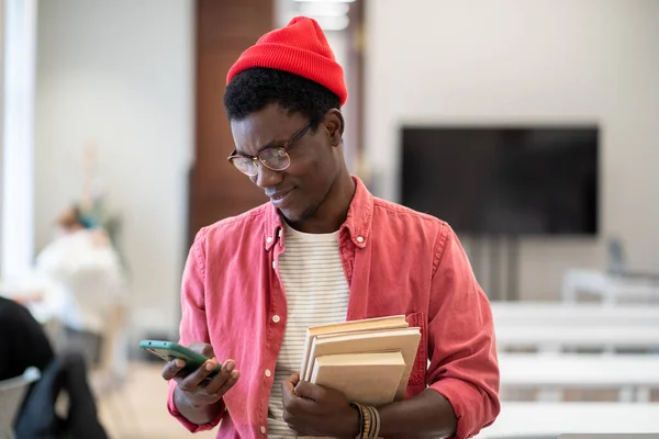 Pleased black student man stands in campus library with textbooks using smartphone on study break. Satisfied African American guy scroll web posts, chat online, surf internet in university classroom